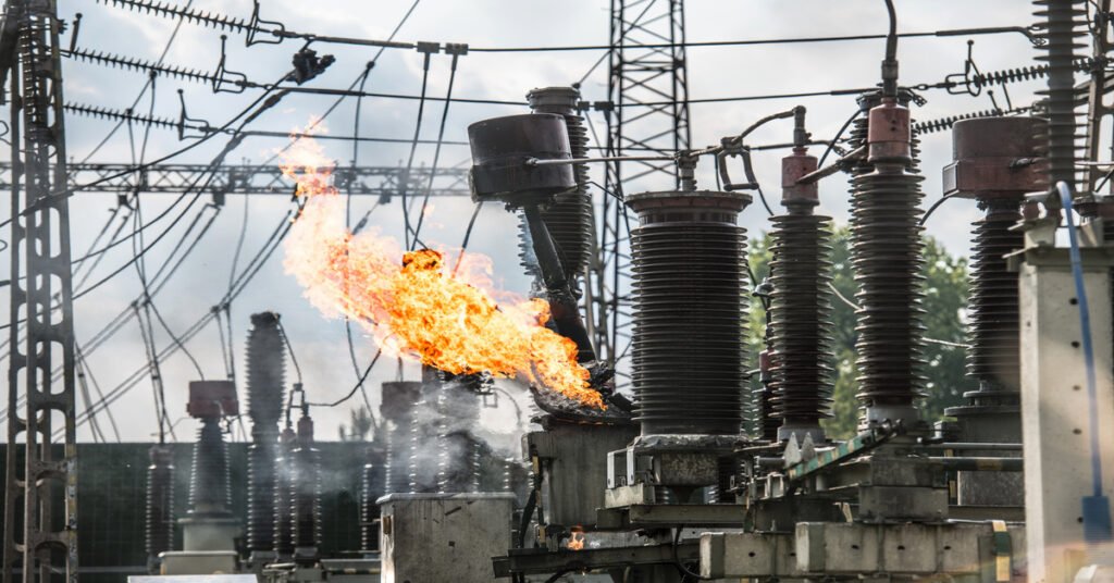 The terminal on an industrial transformer is on fire in a power plant, with other transformers and cables in the background.