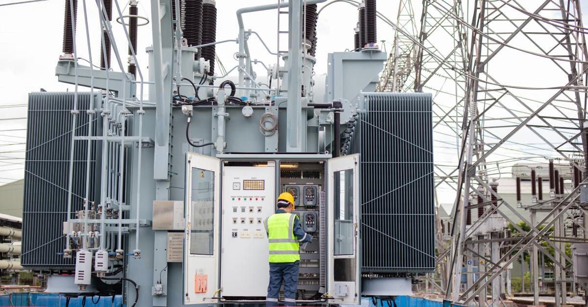 An engineer wearing a safety vest stands in front of a control panel at a large power transformer in a power plant.
