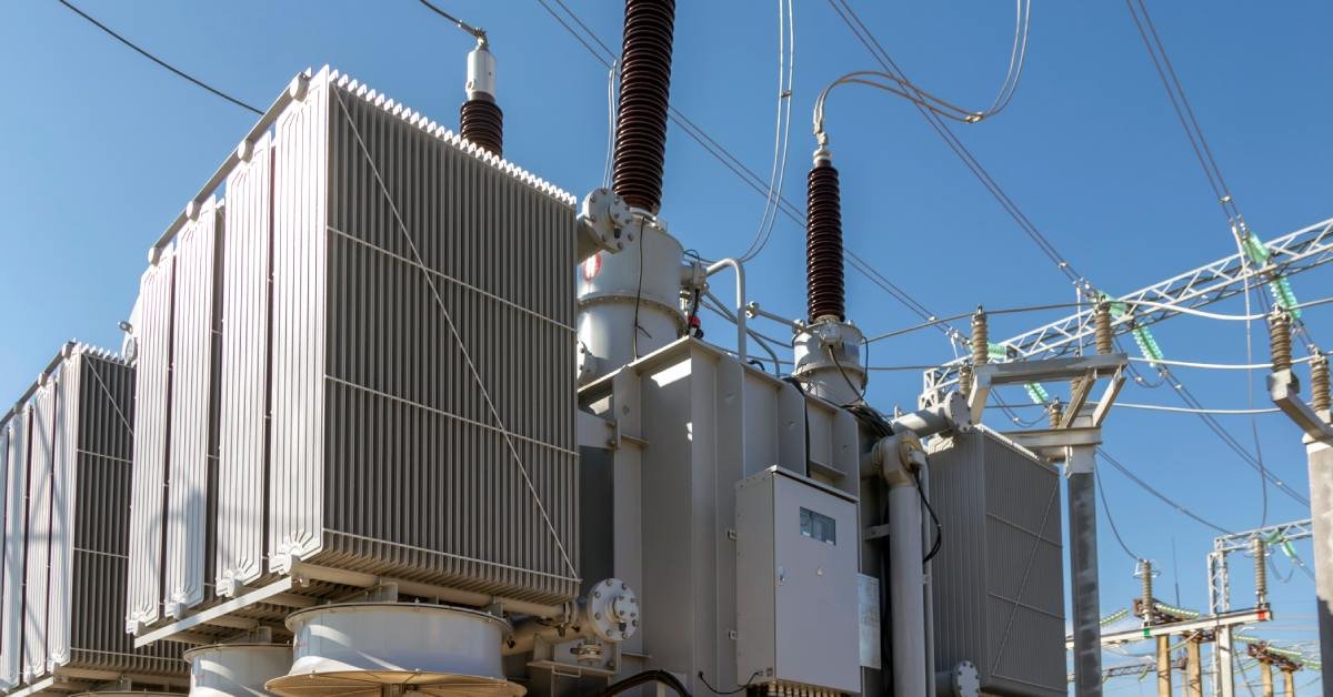 A transformer in a power plant with cooling tubes on each side and three visible terminals connected to hanging cords.