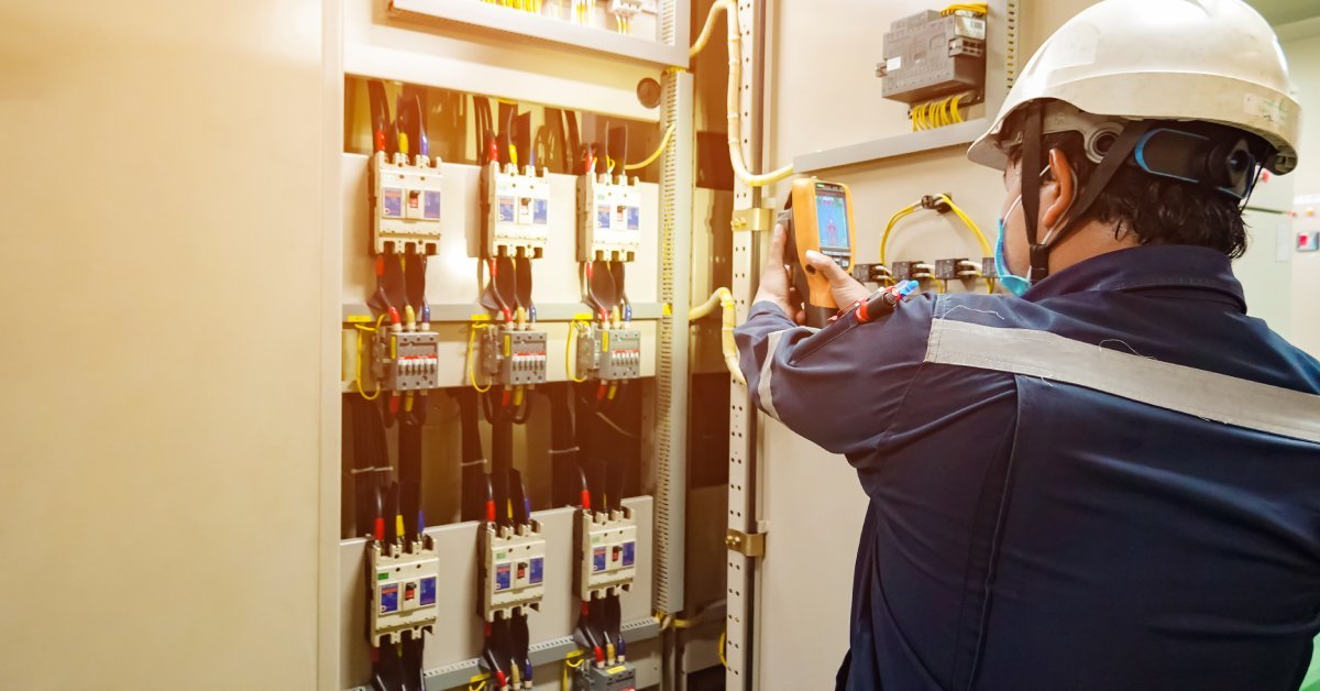 A person wearing a hard hat and a mask uses an infrared laser thermometer to inspect a power transformer located indoors.